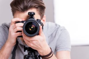 Smile! Young man focusing at you with digital camera while standing in studio with lighting equipment on background